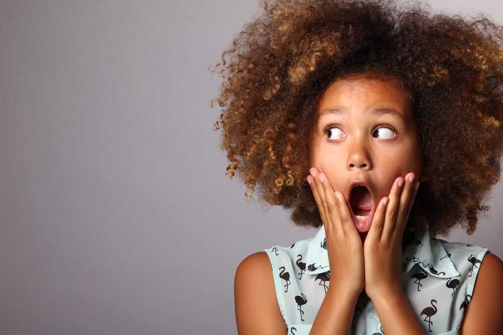 Surprised child with curly hair and open mouth, hands on cheeks against gray background.