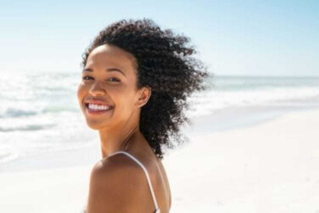 Young woman smiling on a sunny beach with waves in the background.