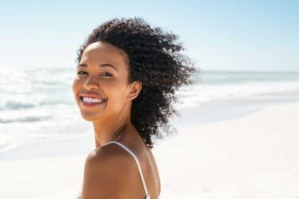 Young woman smiling on a sunny beach with waves in the background.