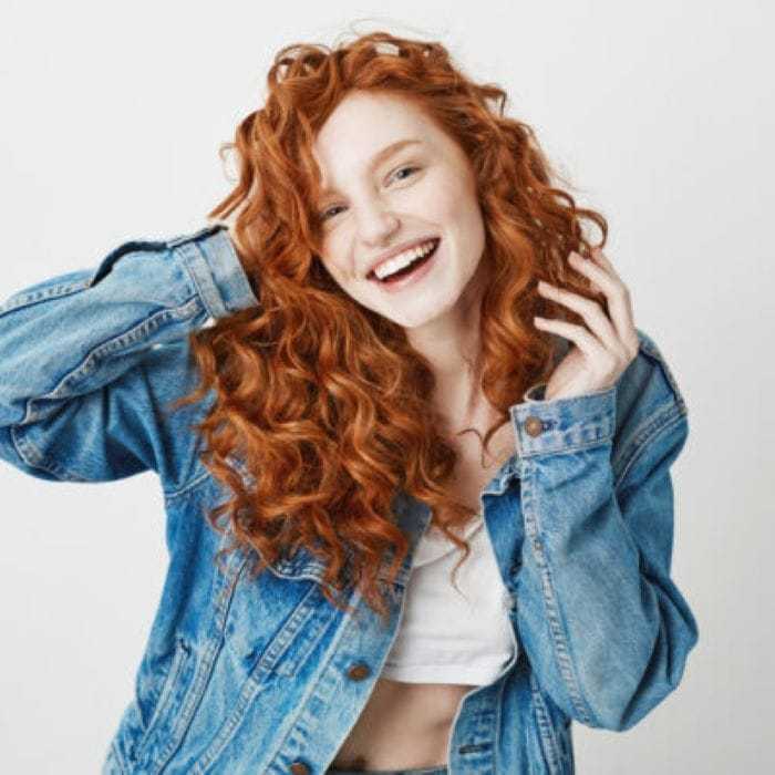 Smiling woman with red curly hair in a denim jacket against a plain white background.