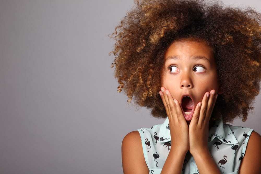 Surprised child with curly hair and open mouth, hands on cheeks against gray background.