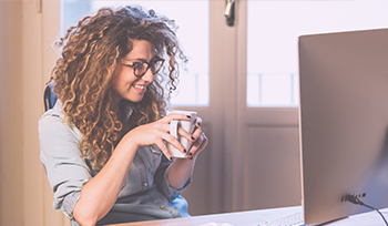 A woman with glasses and curly hair sits at a desk with a cup of coffee smiling at a lesson on her computer screen.